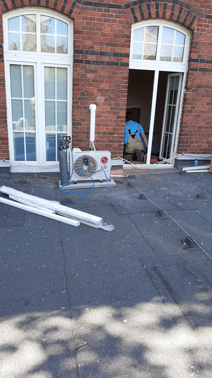 Montaż klimatyzacji na dachu budynku z czerwonej cegły; widoczny agregat LG, butla z gazem i instalator w niebieskiej koszuli w otwartym oknie.