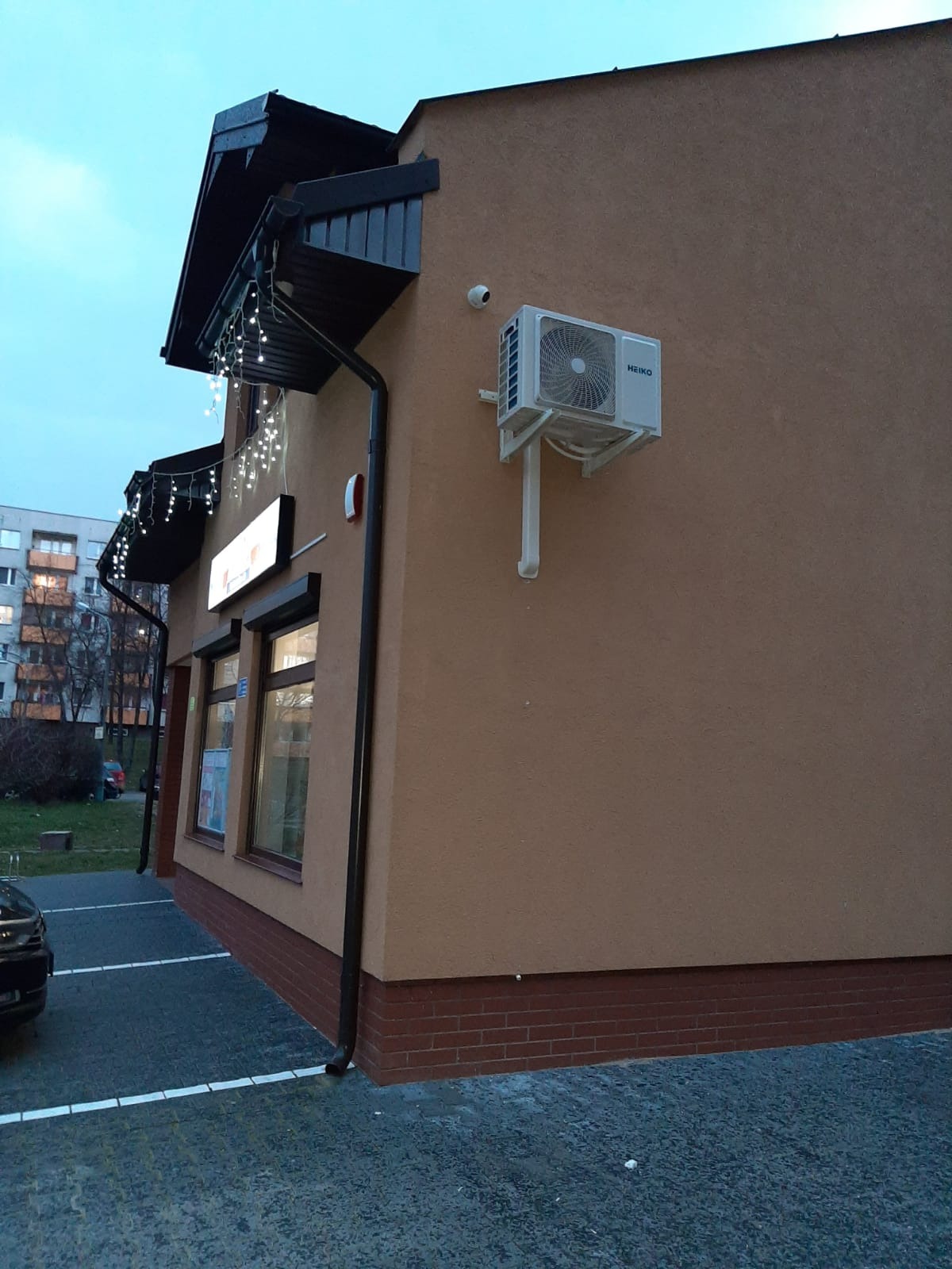 Zewnętrzna jednostka klimatyzacji marki HEIKO zamontowana na ścianie budynku w Radomiu, widoczna rura odprowadzająca skropliny, nad nią kamera monitoringu.
