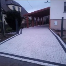 Ułożona jasnoszara kostka brukowa z ciemnoszarym obramowaniem prowadząca do drewnianej wiaty garażowej przy domu.