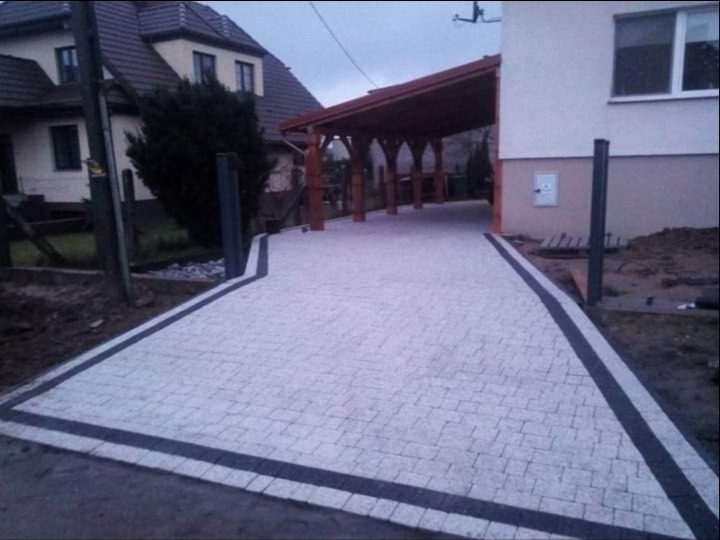 Ułożona jasnoszara kostka brukowa z ciemnoszarym obramowaniem prowadząca do drewnianej wiaty garażowej przy domu.
