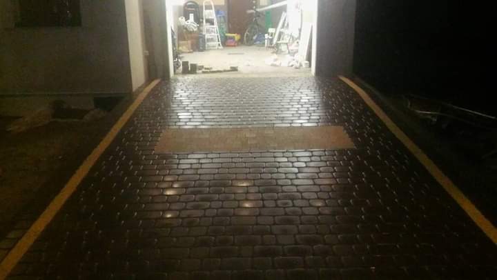 Ułożona, ciemna kostka brukowa prowadząca do garażu z widoczną, jaśniejszą wstawką i żółtym obrzeżem, w tle drabina i rower.