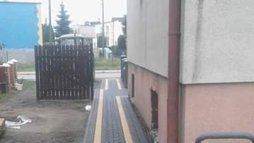 Ułożony chodnik z szarej kostki brukowej z żółtymi paskami, prowadzący od furtki do budynku. Widoczne ślady piasku i niedokończone prace.