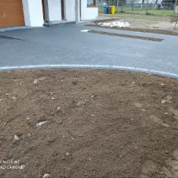 Ułożona szara kostka brukowa na podjeździe domu, obrzeża z jasnoszarego granitu, widoczny fragment rabaty ziemnej przed kostką.