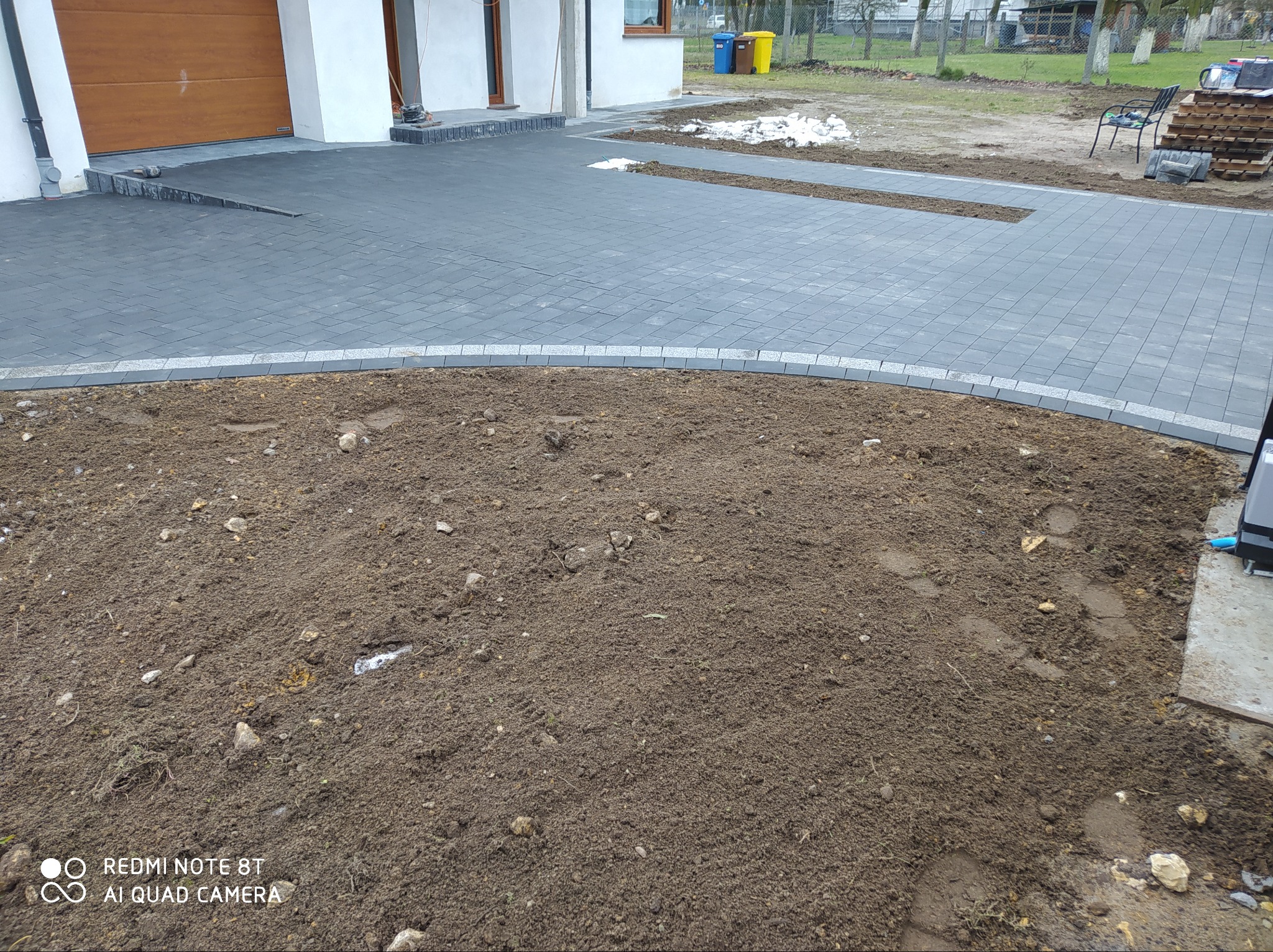 Ułożona szara kostka brukowa na podjeździe domu, obrzeża z jasnoszarego granitu, widoczny fragment rabaty ziemnej przed kostką.