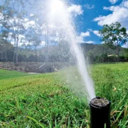 Automatyczny zraszacz trawnikowy w akcji, wyrzucający strumień wody na tle zielonego trawnika, drzew i błękitnego nieba z chmurami.