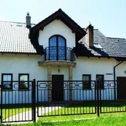 Dom jednorodzinny z ciemną dachówką ceramiczną, balkonem z kutą balustradą i czarnym ogrodzeniem z przodu.