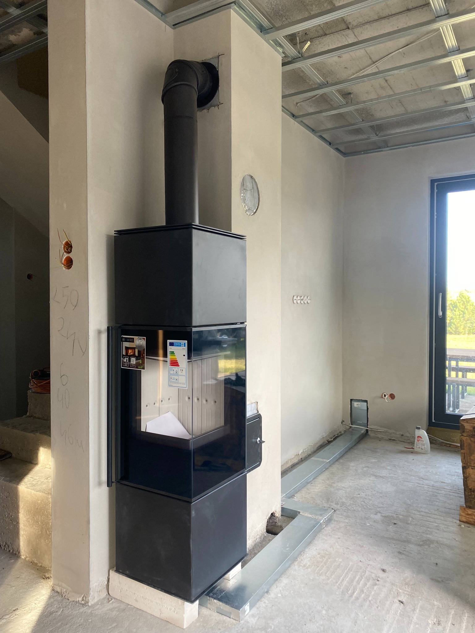 Czarny piec wolnostojący REN z dolotem powietrza, w surowym wnętrzu z widocznymi instalacjami i trójnikiem ceramicznym w kominie systemowym. Widoczne schody i okno.