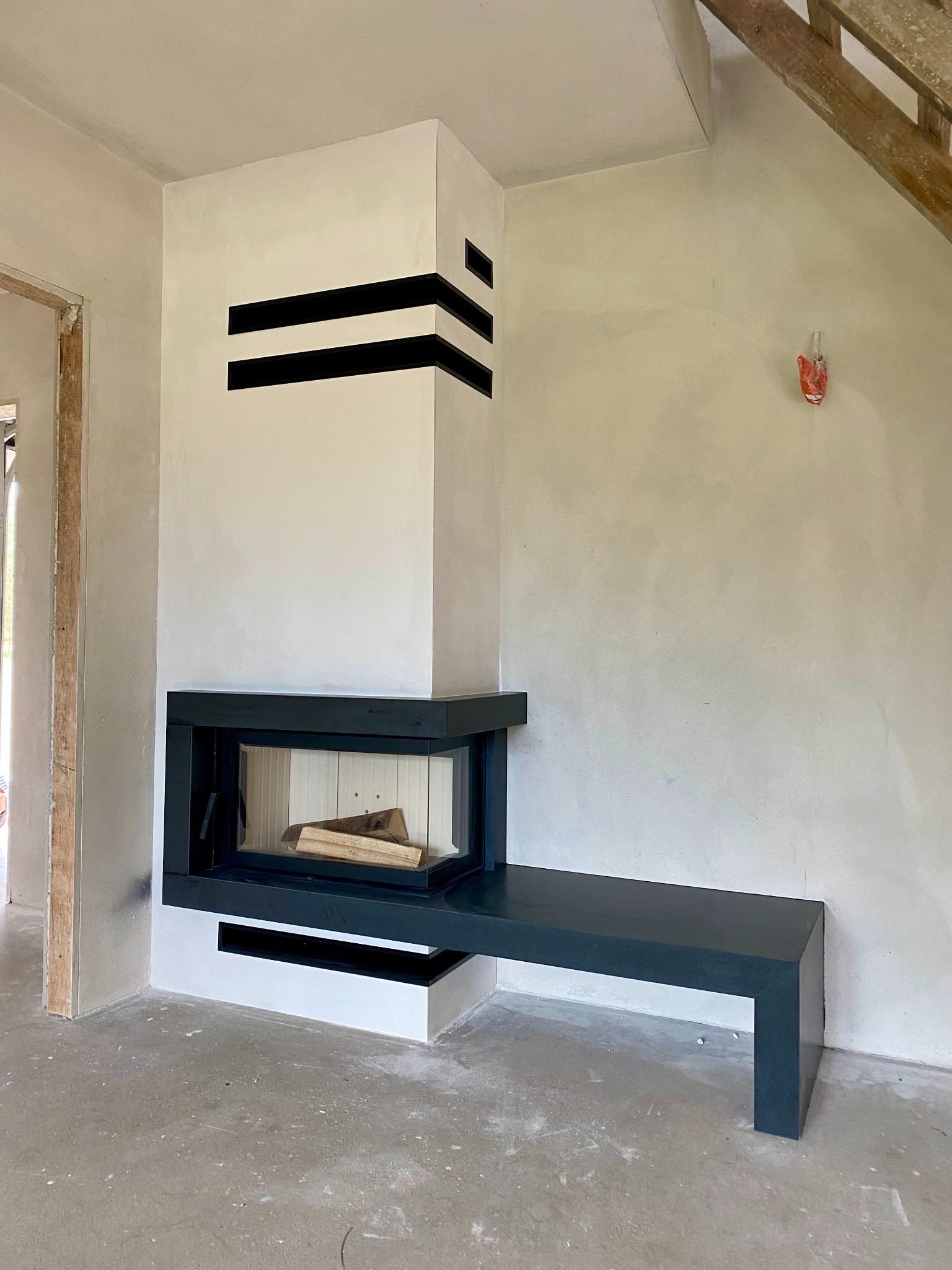 Nowoczesny kominek z czarnymi akcentami i drewnem w palenisku, w surowym wnętrzu z betonową podłogą i schodami. Widoczne niedokończone wykończenia.