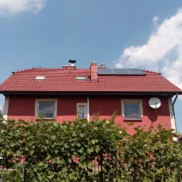 Czerwony dom jednorodzinny z panelami słonecznymi na dachu, widoczne zza zielonego żywopłotu.