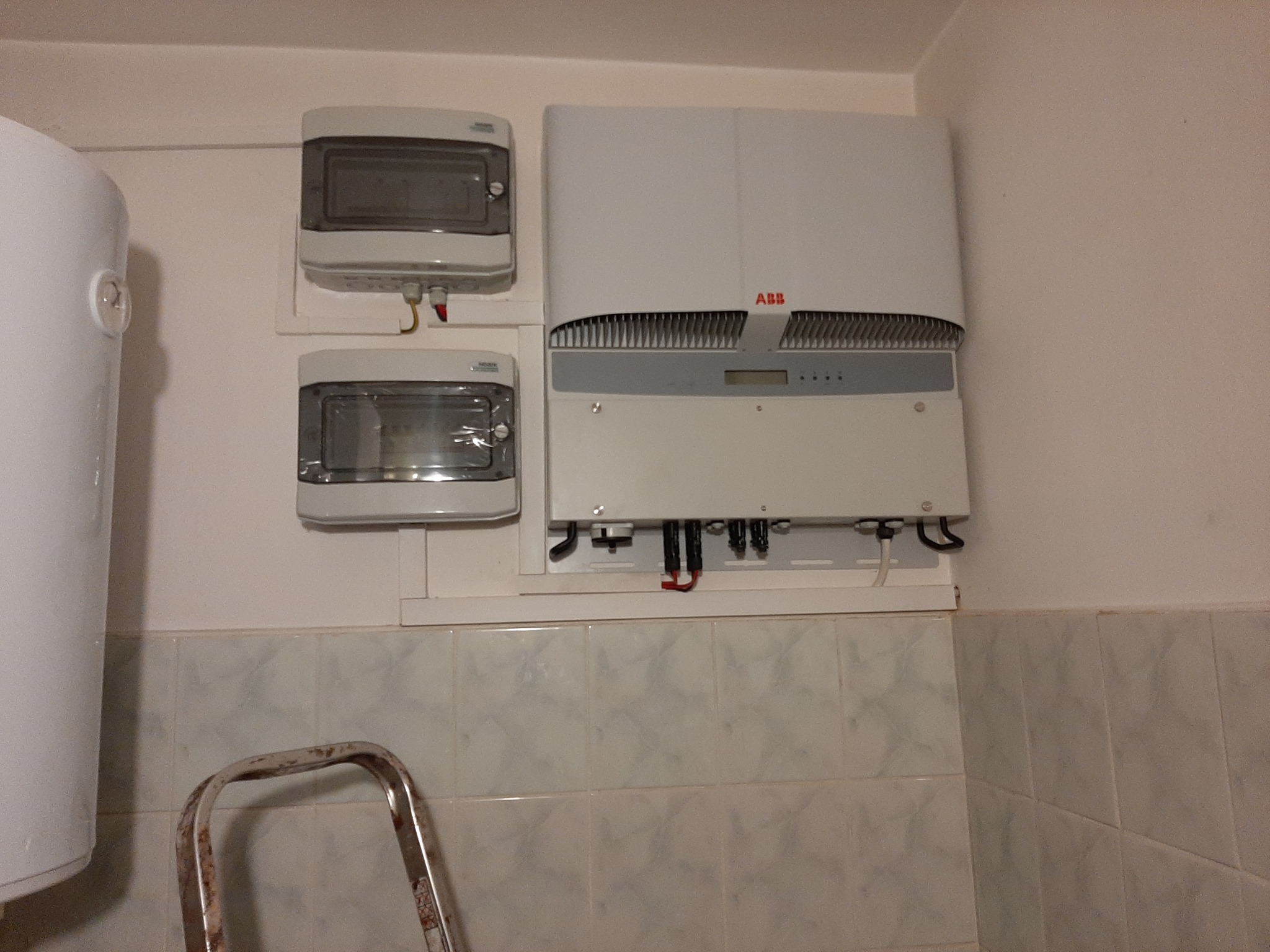 Szara ściana z zamontowanym białym inwerterem ABB, dwoma skrzynkami bezpiecznikowymi i fragmentem drabiny w dolnej części kadru.