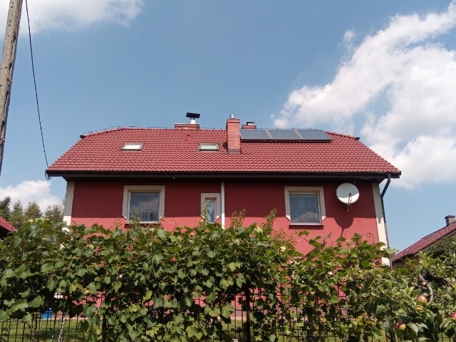 Czerwony dom jednorodzinny z panelami słonecznymi na dachu, widoczne zza zielonego żywopłotu.