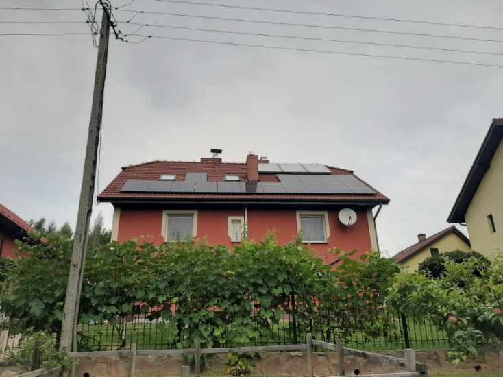 Czerwony dom jednorodzinny z zamontowanymi panelami słonecznymi na dachu, widocznymi pomiędzy oknami dachowymi, antena satelitarna na ścianie bocznej, pochmurne niebo.