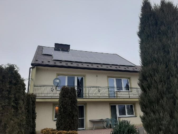 Dwupiętrowy dom jednorodzinny z panelami słonecznymi na dachu pokrytym brązową blachodachówką, widok z poziomu gruntu w pochmurny dzień.