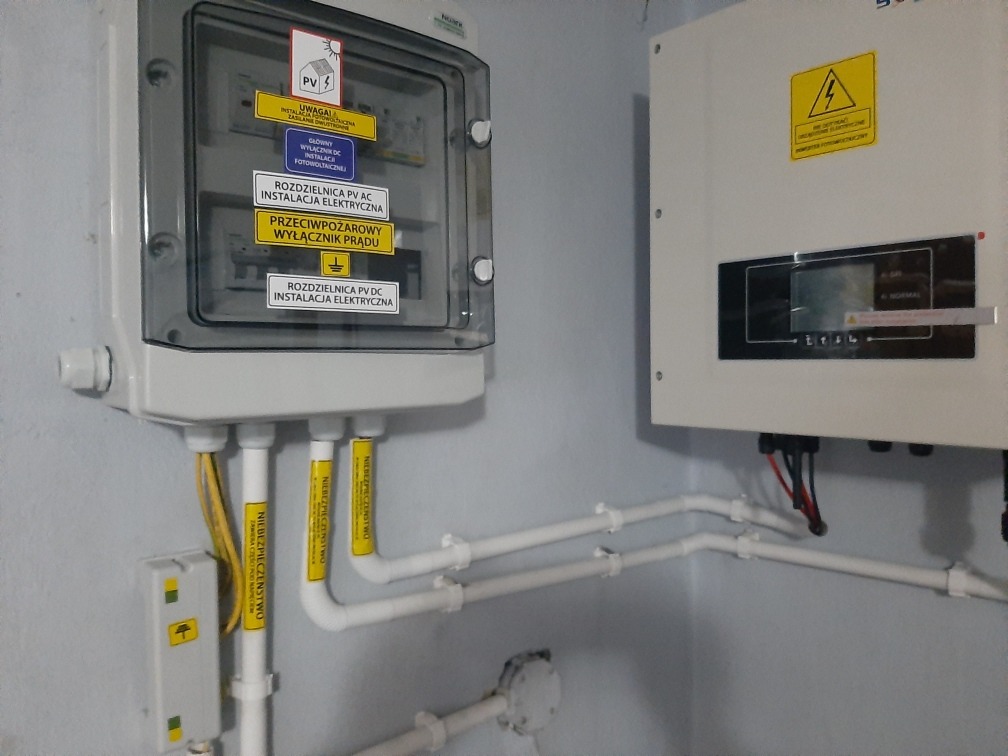 Rozdzielnica AC/DC i falownik instalacji, z widocznymi etykietami ostrzegawczymi i przewodami w rurach PCV, zamontowane na ścianie w pomieszczeniu.