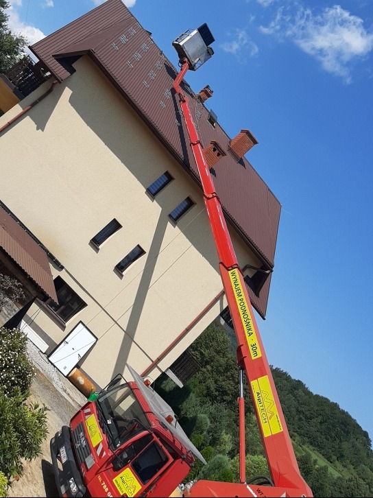 Montaż paneli fotowoltaicznych na dachu dwuspadowym z wykorzystaniem podnośnika koszowego na tle błękitnego nieba.