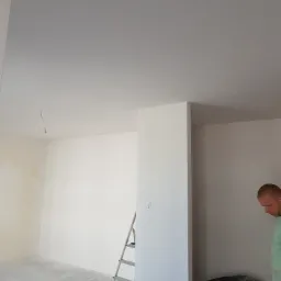 Wnętrze w trakcie prac, ściany i sufit pomalowane na biało, widoczna drabina aluminiowa, przewody wystające z sufitu i malarz w tle.