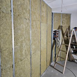 Roboty ogólnobudowlane Kamil Kalisz - Montaż izolacji z wełny mineralnej w metalowym stelażu ściany działowej, widoczna drabina i instalacja elektryczna w trakcie prac.