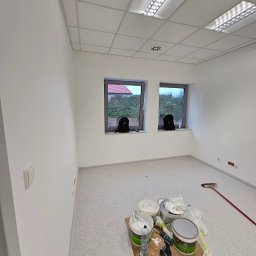 Roboty ogólnobudowlane Kamil Kalisz - Pomalowane na biało wnętrze biura z dwoma oknami, na podłodze karton z puszkami farb, mieszadłem i ścierką, widoczne lampy rastrowe w suficie podwieszanym.