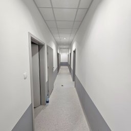 Roboty ogólnobudowlane Kamil Kalisz - Długi, pusty korytarz biurowy po odświeżeniu, z pomalowanymi na biało ścianami i szarym pasem przy podłodze, sufit kasetonowy, widoczna butelka wody przy futrynie.