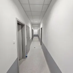 Długi, pusty korytarz biurowy po odświeżeniu, z pomalowanymi na biało ścianami i szarym pasem przy podłodze, sufit kasetonowy, widoczna butelka wody przy futrynie.