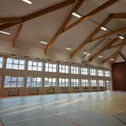 Wnętrze hali sportowej z drewnianą konstrukcją dachu, rzędem okien i szarą podłogą z liniami boiska, widoczne ślady stóp.