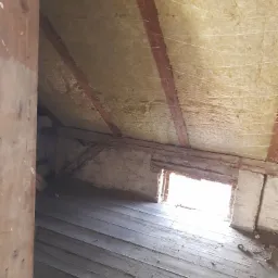 Poddasze przed remontem: widok na ocieplony dach z widocznymi belkami, surową podłogę z desek i okno w szczycie ściany.