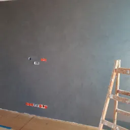 Ściana pokryta szarą dekoracyjną powłoką z widocznymi puszkami elektrycznymi, zabezpieczona podłoga i drewniana drabina malarska.