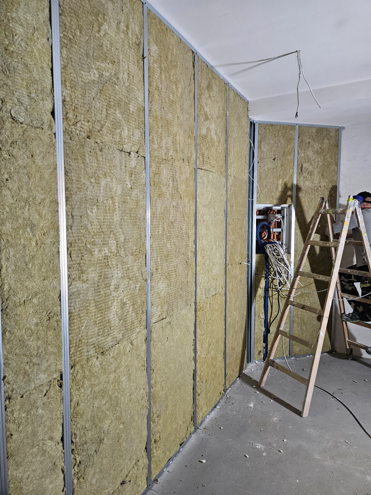 Montaż izolacji z wełny mineralnej w metalowym stelażu ściany działowej, widoczna drabina i instalacja elektryczna w trakcie prac.