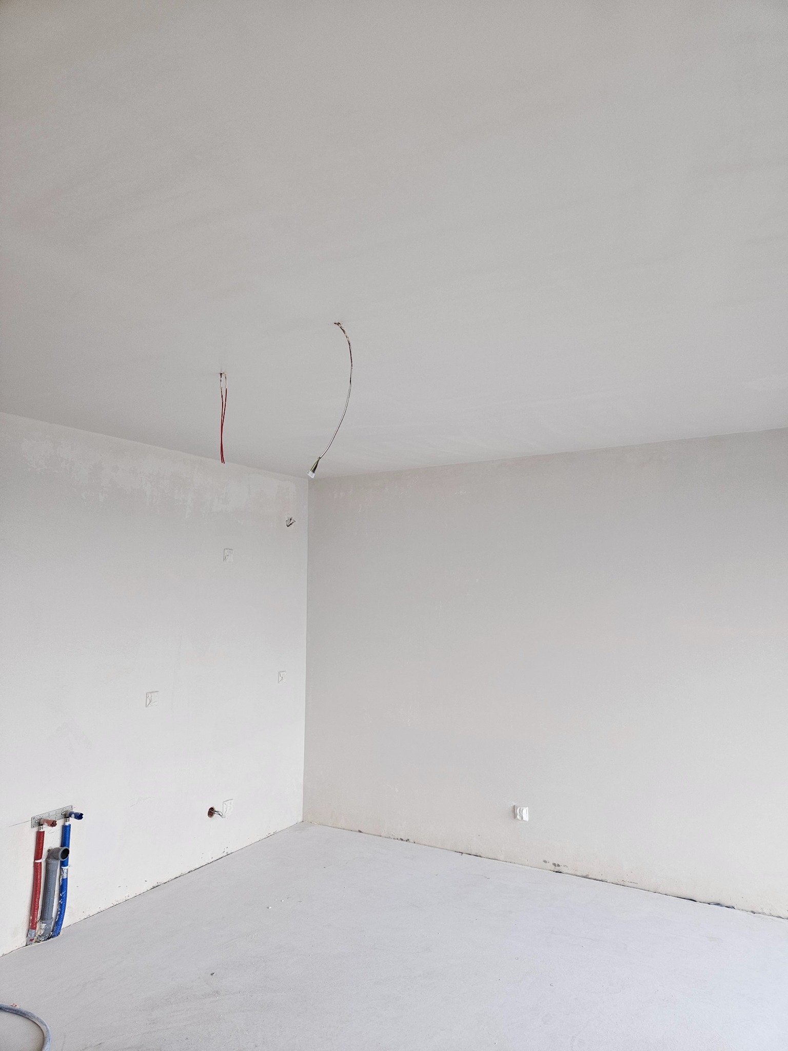 Surowe wnętrze w trakcie prac: ściany po wstępnym wyrównaniu, widoczne instalacje elektryczne i hydrauliczne, podłoga przygotowana pod wylewkę, brak wykończenia.