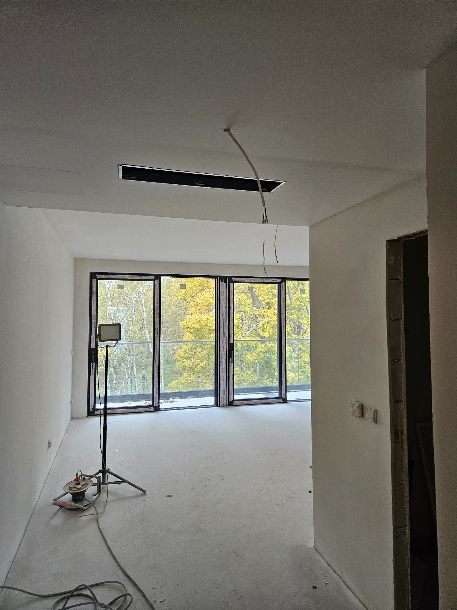 Wnętrze w trakcie prac wykończeniowych: ściany przygotowane pod malowanie, widoczna szlifierka do gładzi na posadzce, na zewnątrz jesienny krajobraz.