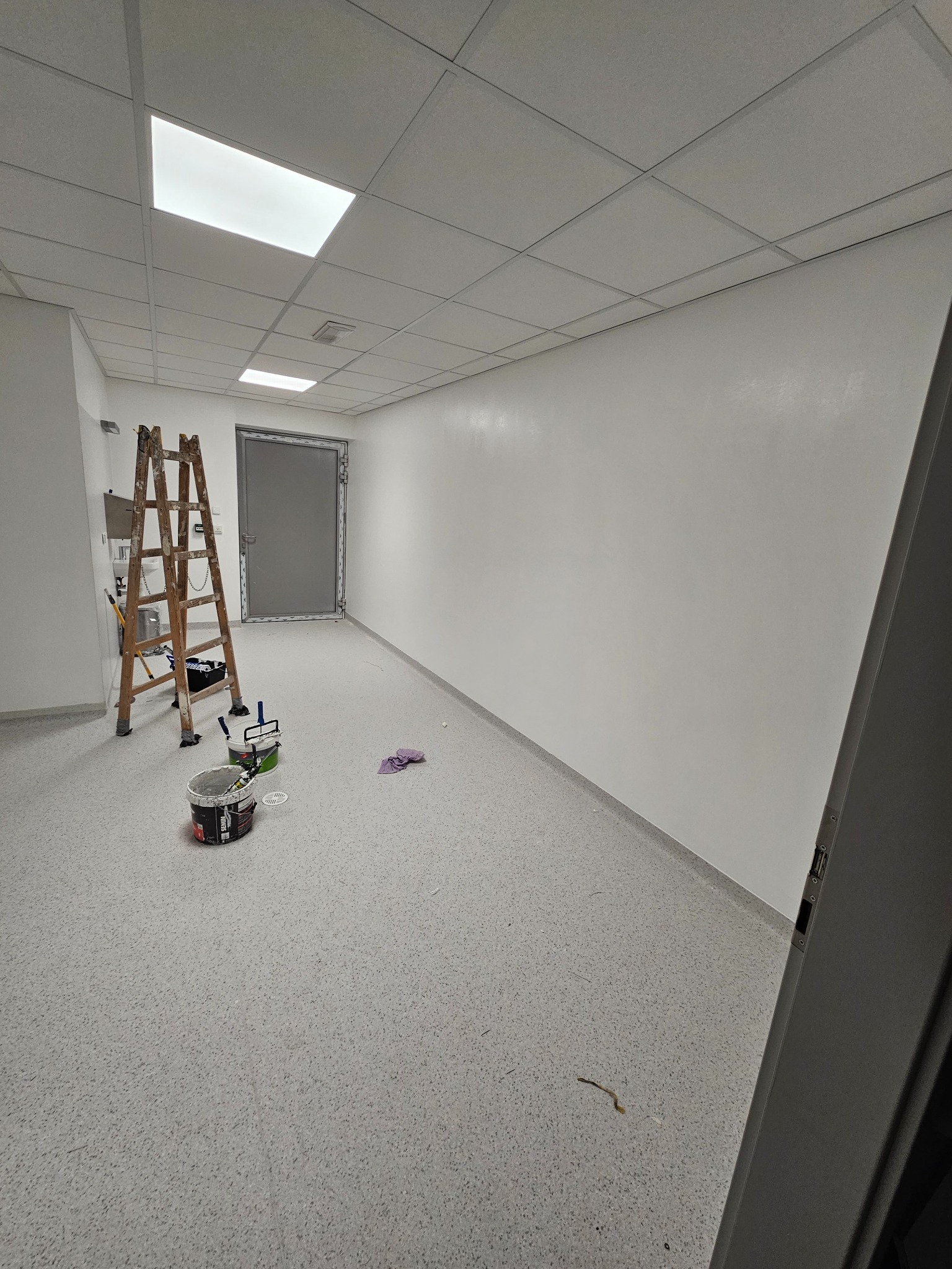 Wnętrze biurowe w trakcie odświeżania: drewniana drabina oparta o ścianę, puszki z farbą, wałki malarskie, ślady świeżego malowania na ścianie, sufit kasetonowy z oświetleniem LED.