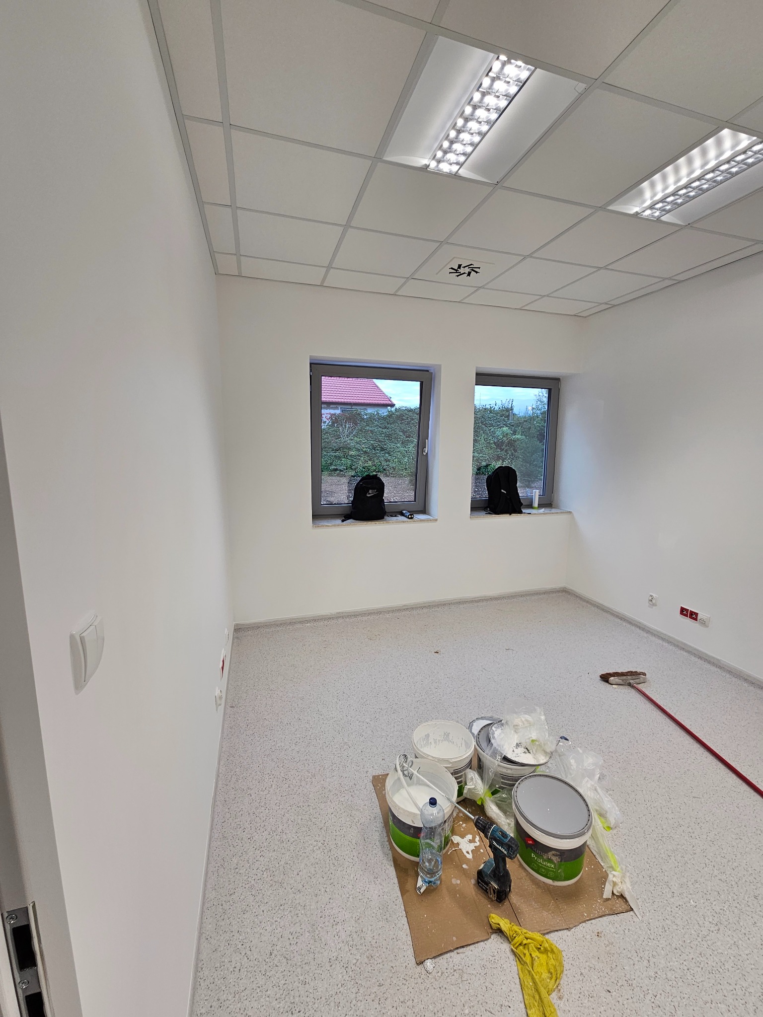 Pomalowane na biało wnętrze biura z dwoma oknami, na podłodze karton z puszkami farb, mieszadłem i ścierką, widoczne lampy rastrowe w suficie podwieszanym.