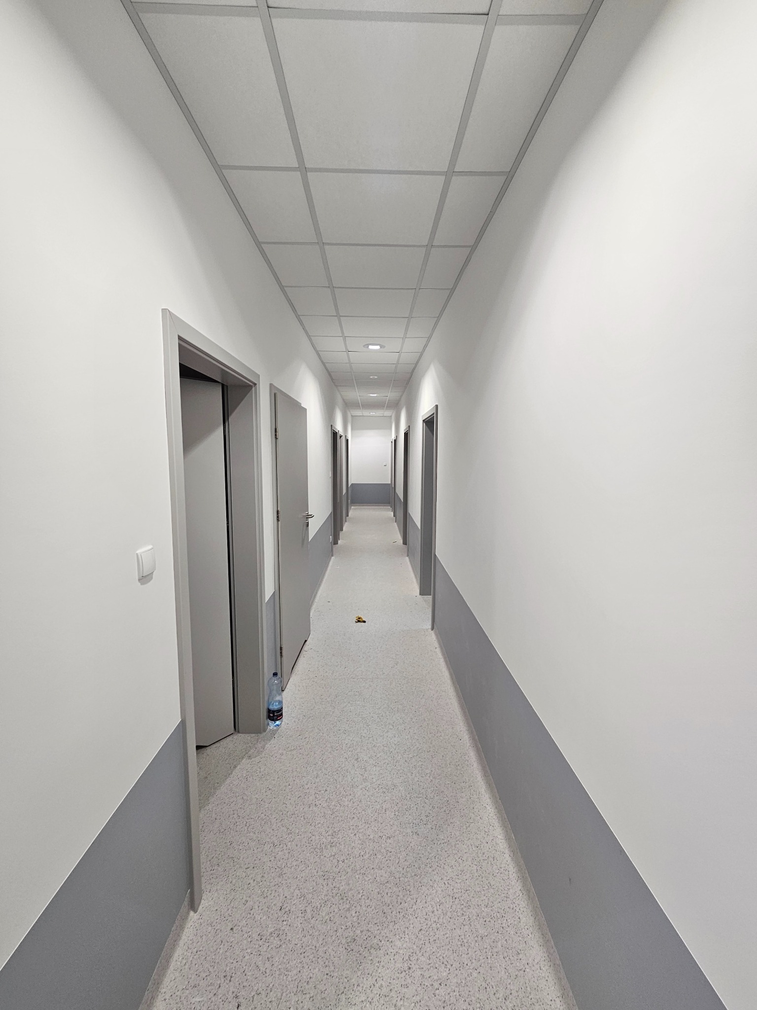 Długi, pusty korytarz biurowy po odświeżeniu, z pomalowanymi na biało ścianami i szarym pasem przy podłodze, sufit kasetonowy, widoczna butelka wody przy futrynie.