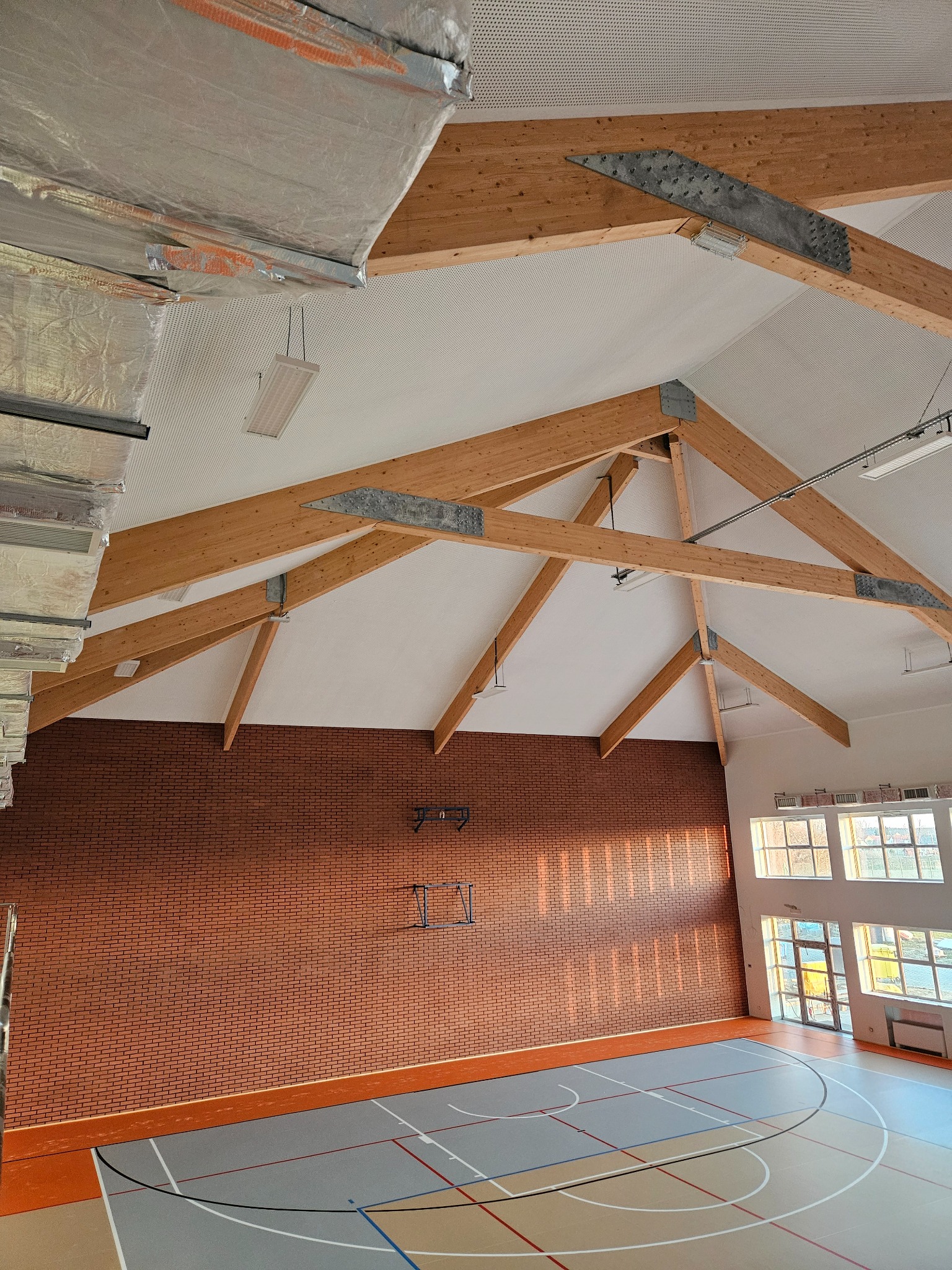 Wysokie wnętrze hali sportowej z drewnianą konstrukcją dachu, białym perforowanym sufitem podwieszanym i ceglaną ścianą, widoczne elementy instalacji wentylacyjnej.