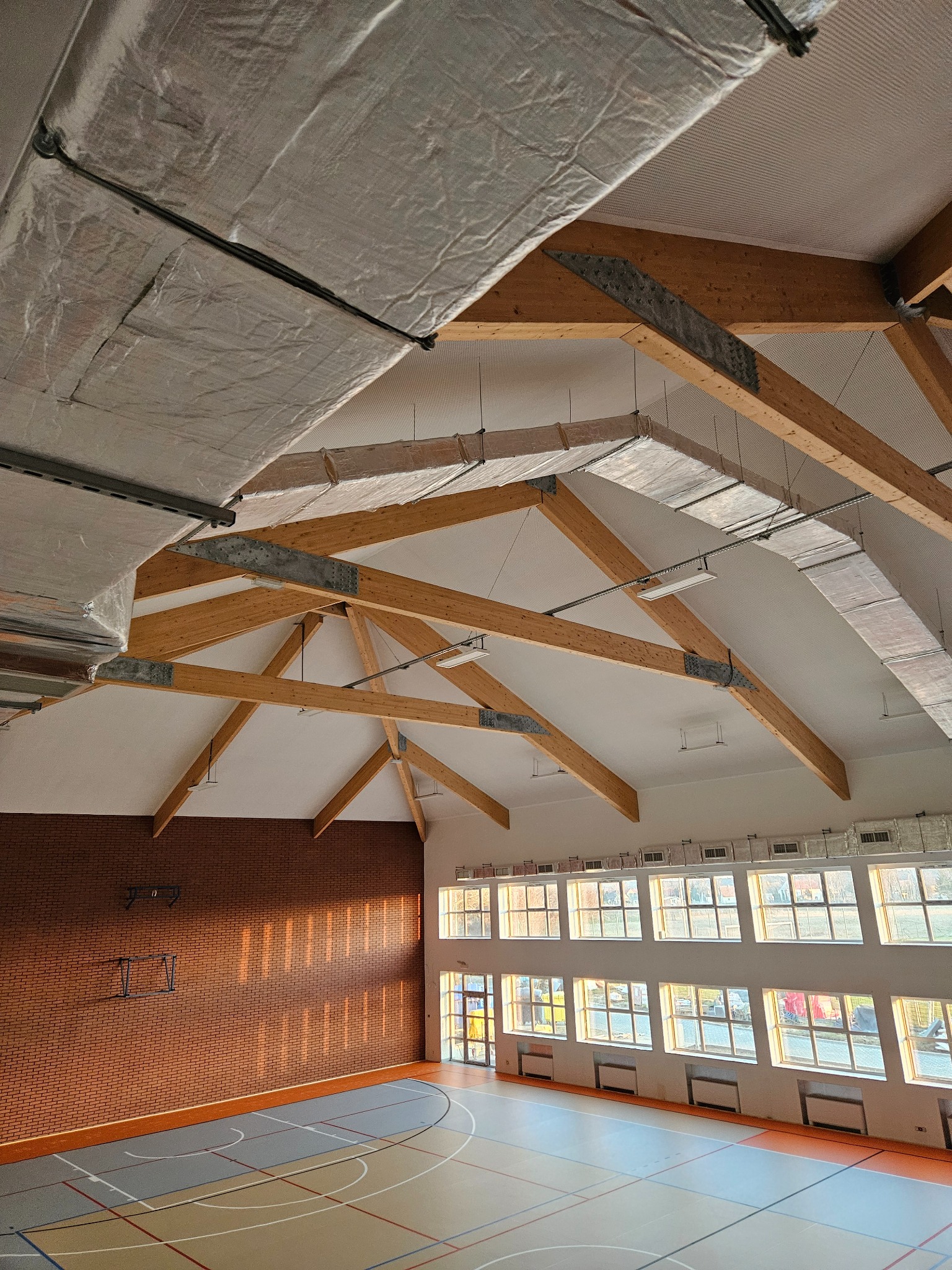 Wysokie wnętrze sali gimnastycznej z drewnianą konstrukcją dachu i oknami na całej ścianie, widoczne instalacje na suficie.