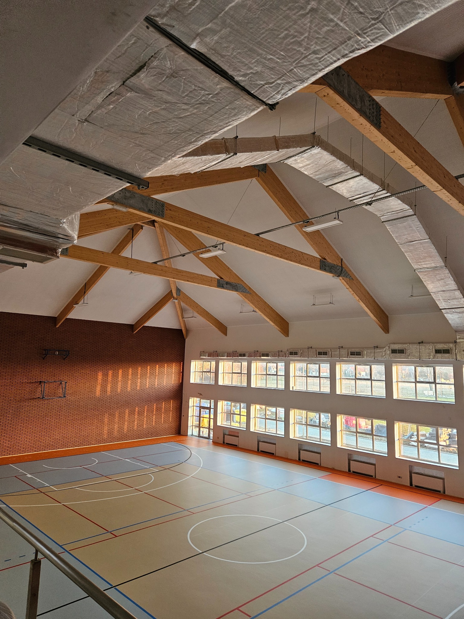 Wysokie wnętrze hali sportowej z drewnianą konstrukcją dachu, ceglaną ścianą boczną i parkietem w linie boisk, widoczne kanały wentylacyjne pod sufitem.