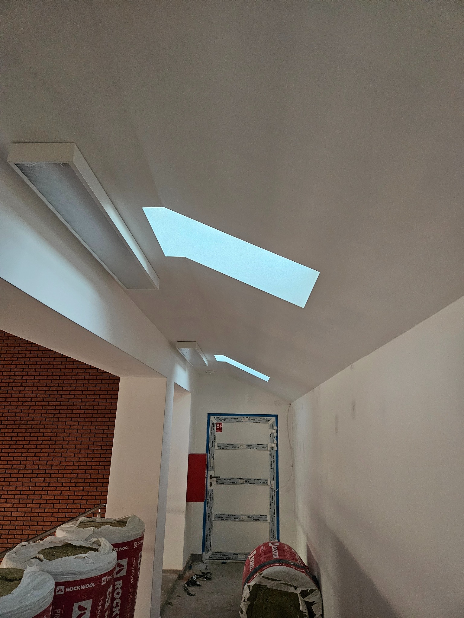 Wnętrze w trakcie prac wykończeniowych: widoczne worki z wełną mineralną Rockwool, sufit z oświetleniem w kształcie prostokątów, ściany przygotowane pod malowanie, drzwi z białymi framugami.