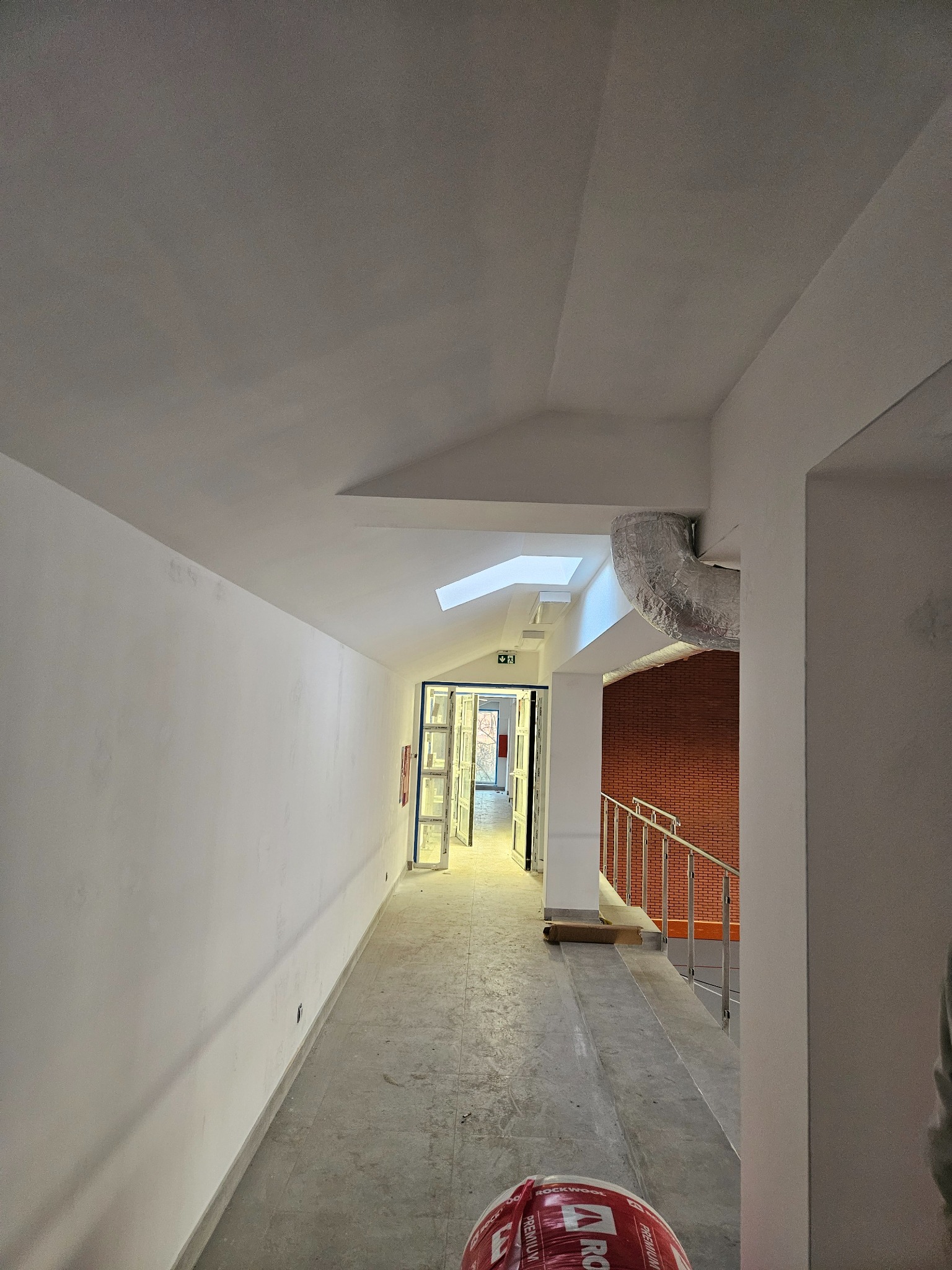 Korytarz w budynku w trakcie prac wykończeniowych, widoczne białe ściany z niedoskonałościami, betonowa podłoga, instalacja wentylacyjna pod sufitem i materiały budowlane w tle.