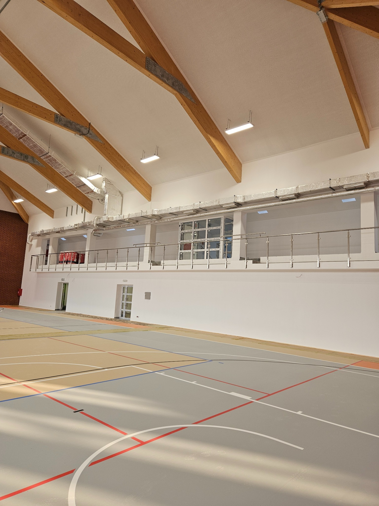 Wnętrze hali sportowej z drewnianą konstrukcją dachu, białymi ścianami i szarą podłogą z kolorowymi liniami boiska.