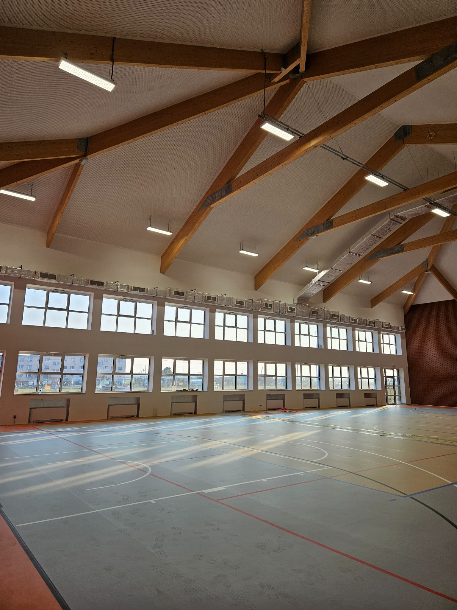 Wnętrze hali sportowej z drewnianą konstrukcją dachu, rzędem okien i szarą podłogą z liniami boiska, widoczne ślady stóp.