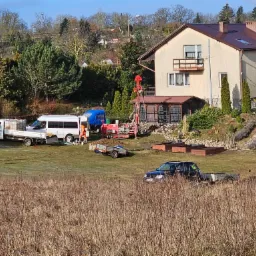 Wiercenie na posesji z domem w tle: wiertnica, ekipa w pomarańczowych kombinezonach, ciężarówki i przyczepka na trawniku.