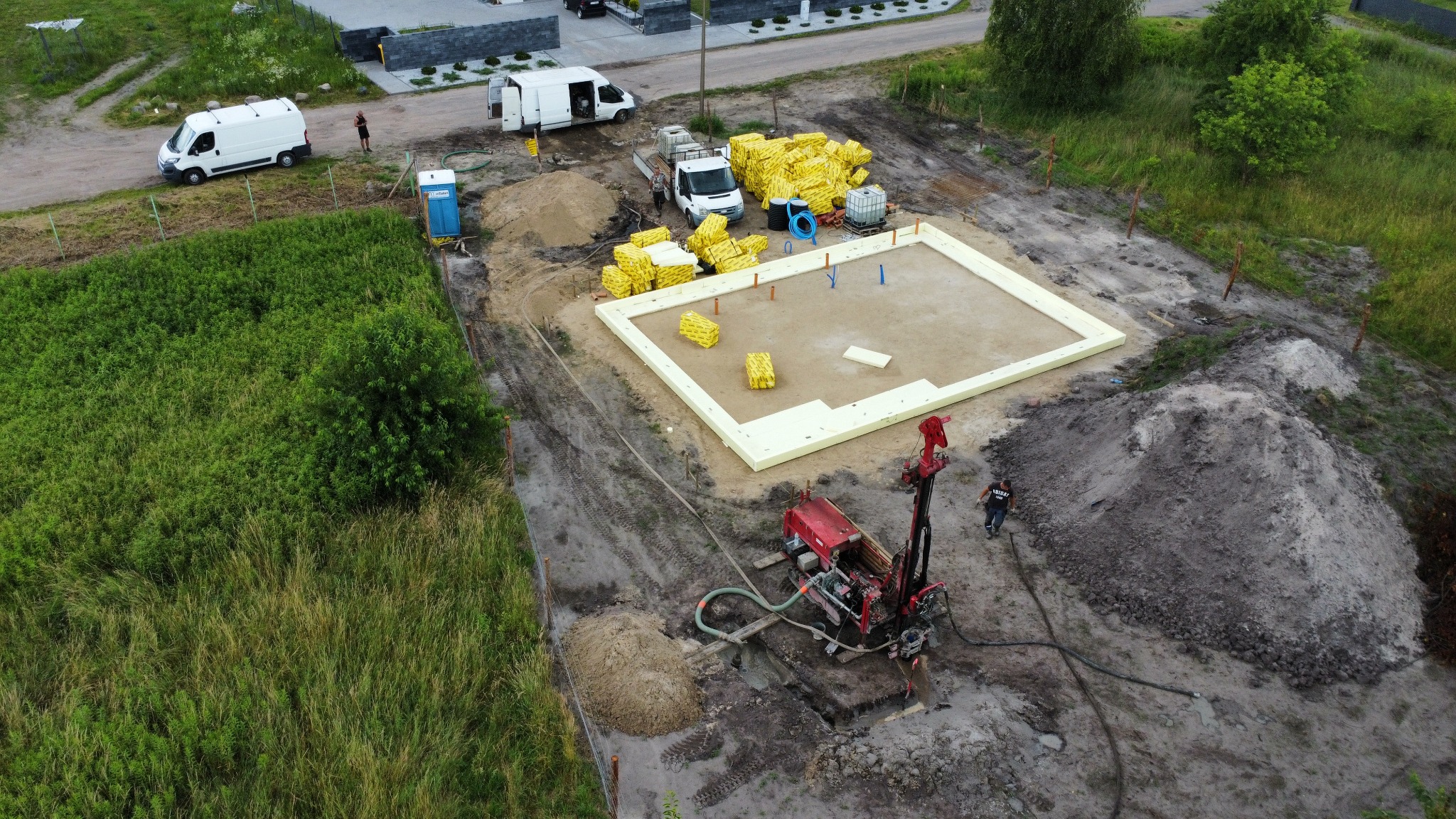Z lotu ptaka widok placu budowy z czerwoną wiertnicą, fundamentami domu z żółtym ociepleniem, stertami materiałów budowlanych, przenośną toaletą i dwoma białymi samochodami dostawczymi.