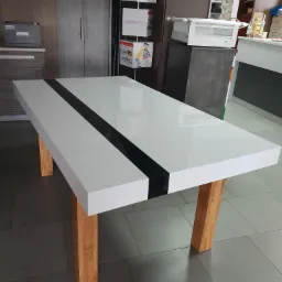 Biały stół z połyskiem i czarnym pasem pośrodku, na drewnianych nogach, widok z góry, w tle zabudowa meblowa.