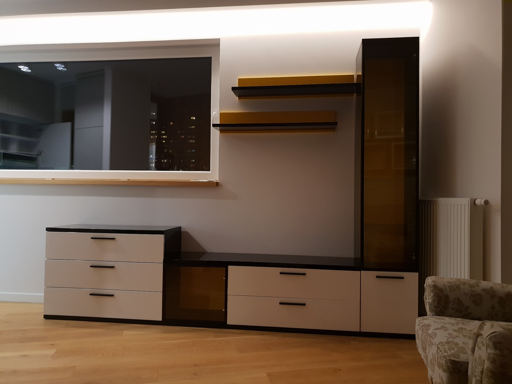 Nowoczesna zabudowa meblowa w salonie z białymi frontami i czarnymi blatami, uzupełniona o półki w odcieniach brązu i przeszkloną witrynę, widoczna na tle okna z nocnym widokiem miasta.