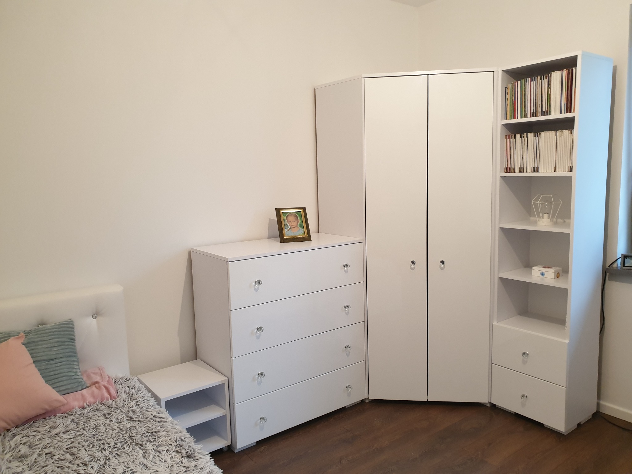 Biały komplet mebli do sypialni w Małkini Górnej: szafa narożna z półkami i szafką, komoda z kryształowymi gałkami oraz stolik nocny, ustawione na drewnianej podłodze.