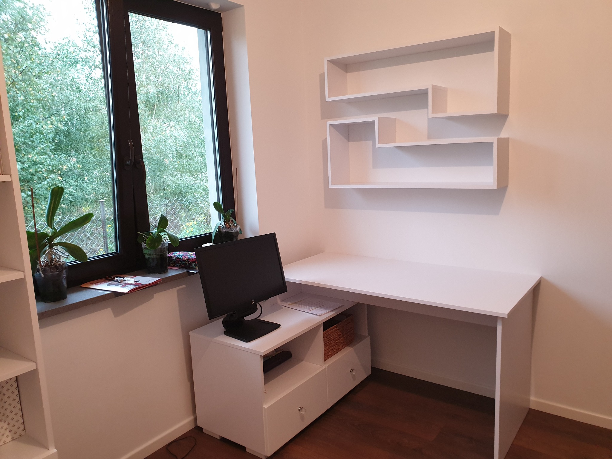 Białe biurko narożne z szafką pod monitor i wiszącymi półkami o nieregularnym kształcie, ustawione przy oknie z widokiem na zieleń.