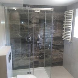 Uslugi Remontowo Budowlane - Nowoczesna łazienka z kabiną prysznicową ze szklanymi drzwiami przesuwnymi, marmurowe płytki na ścianach w odcieniach szarości, biały grzejnik drabinkowy i podwieszana toaleta.