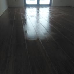 Uslugi Remontowo Budowlane - Świeżo położona, ciemnobrązowa podłoga z paneli, odbijająca światło z okna balkonowego, w surowym, białym pomieszczeniu.