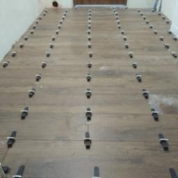 Uslugi Remontowo Budowlane - Układanie brązowych płytek podłogowych imitujących drewno, z użyciem czarnych klipsów i białych klinów poziomujących, widoczne gniazdko elektryczne w ścianie.