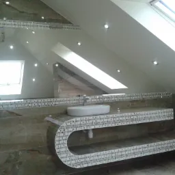 Nowoczesna łazienka na poddaszu z marmurową ścianą i blatem z mozaiki, okrągła umywalka nablatowa, oświetlenie punktowe w skosach sufitu.